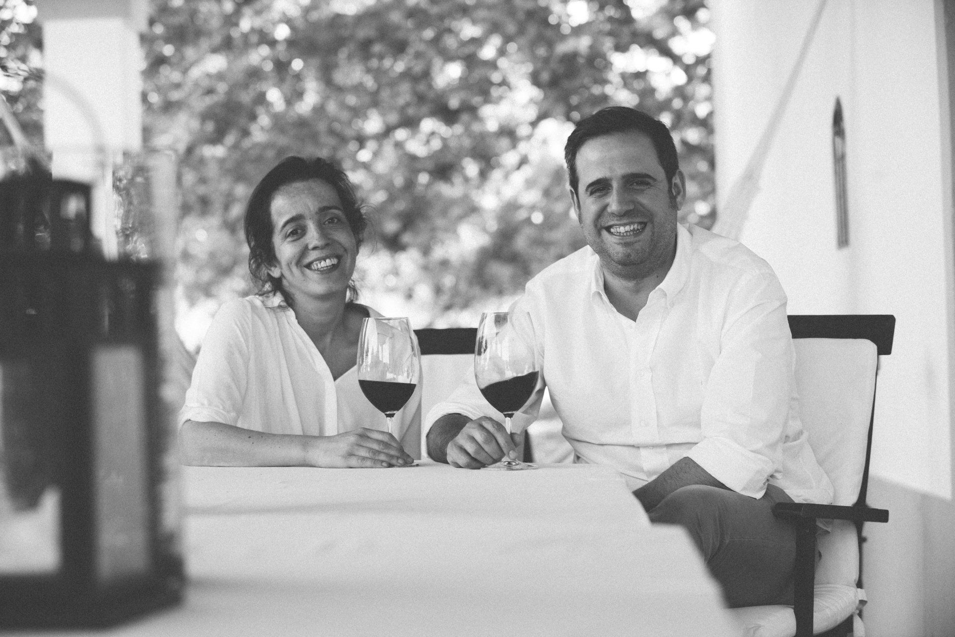 Two people smiling, sitting at a table with red wine glasses. Outdoors, under a white awning.