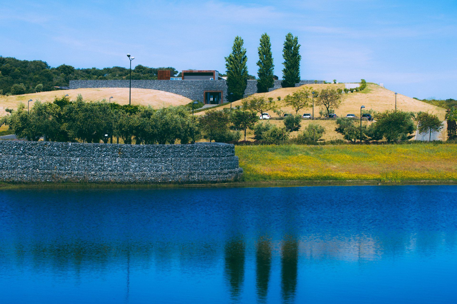 Blue lake with a stone wall and building on a hill under a bright blue sky. Three tall trees stand near the building.