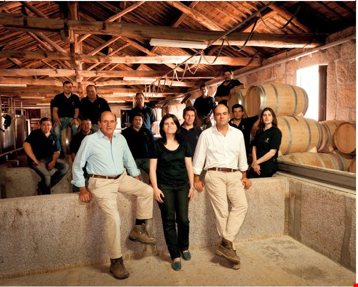 Group of people in a winery; standing near barrels. Sunlight pours in, illuminating wooden rafters and stone walls.