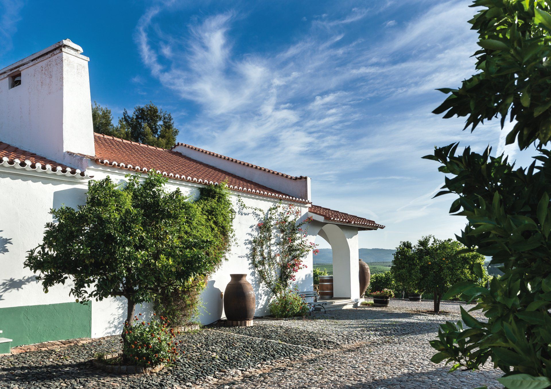 White stucco building with red tile roof under a blue sky, trees in the foreground, gravel path leading to the entrance.