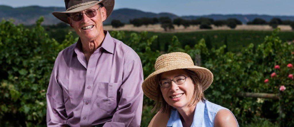 Two people smiling in front of a vineyard, both wearing hats.