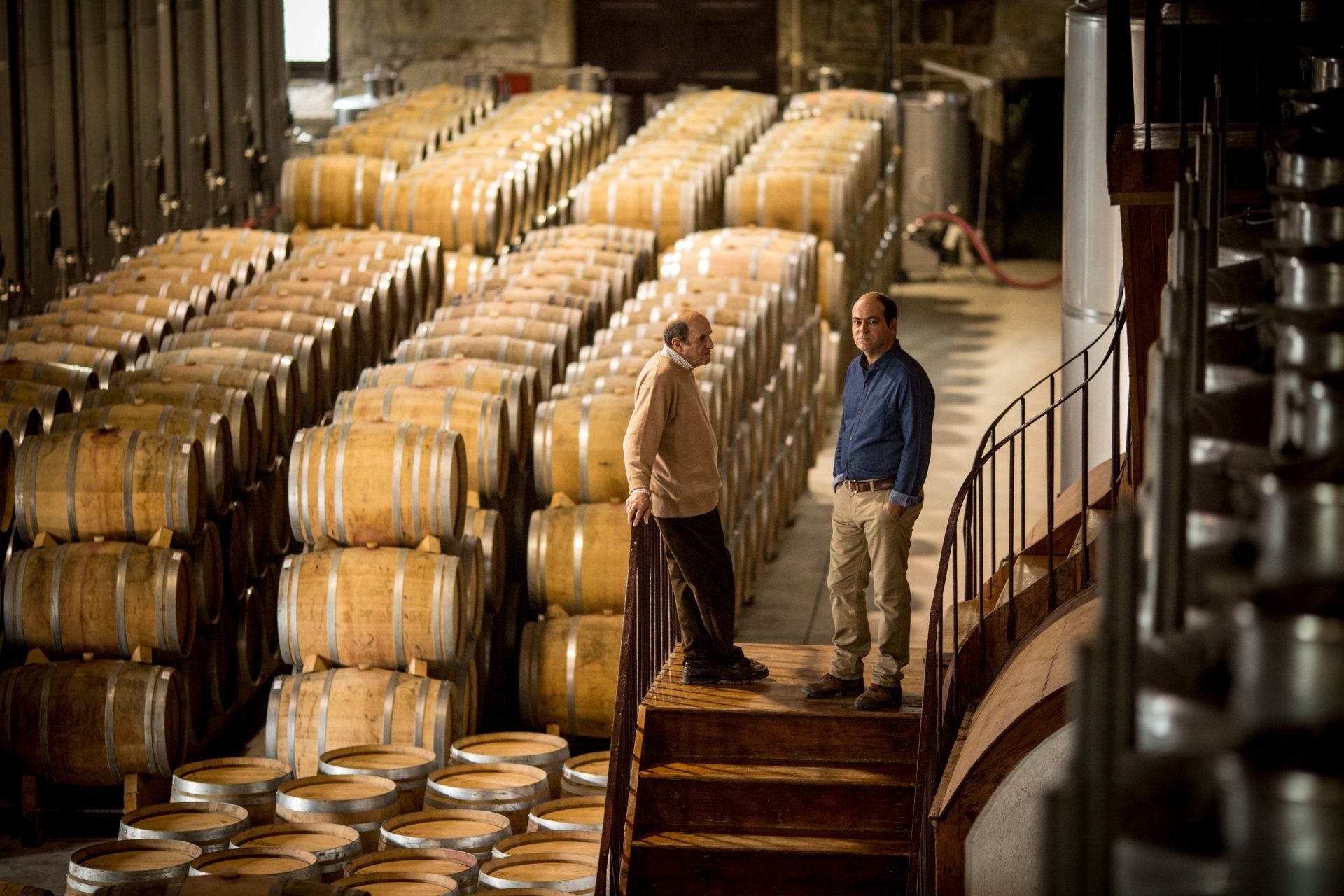 Two men standing on stairs in a cellar filled with stacked wooden barrels.