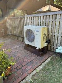 An off-white outdoor mini-split heat pump unit sitting on wooden risers on a brick patio next to a wooden fence.