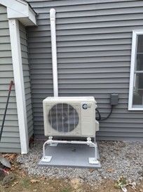 A beige outdoor HVAC heat pump unit mounted on a gray stand against the light gray vinyl siding of a house.