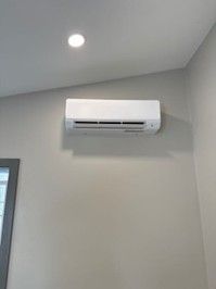 A white wall-mounted air conditioning unit installed on a plain, light gray wall near a ceiling spotlight.