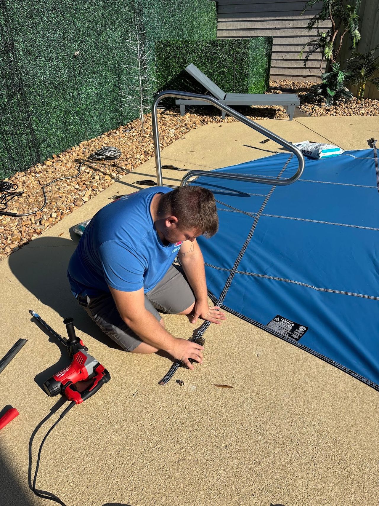 Man installing straps on a blue canopy frame in a gravel yard.