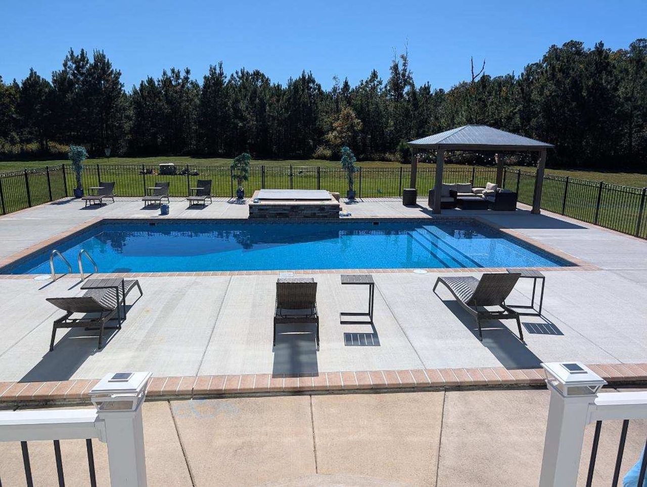 Outdoor pool with lounge chairs, cabanas, and a green landscaped backdrop under a clear blue sky