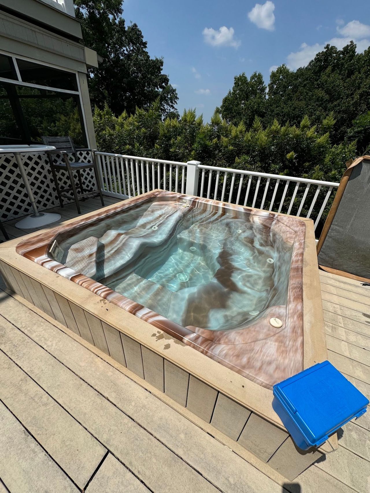 Outdoor hot tub with cover, surrounded by a wooden deck and white railing under trees.