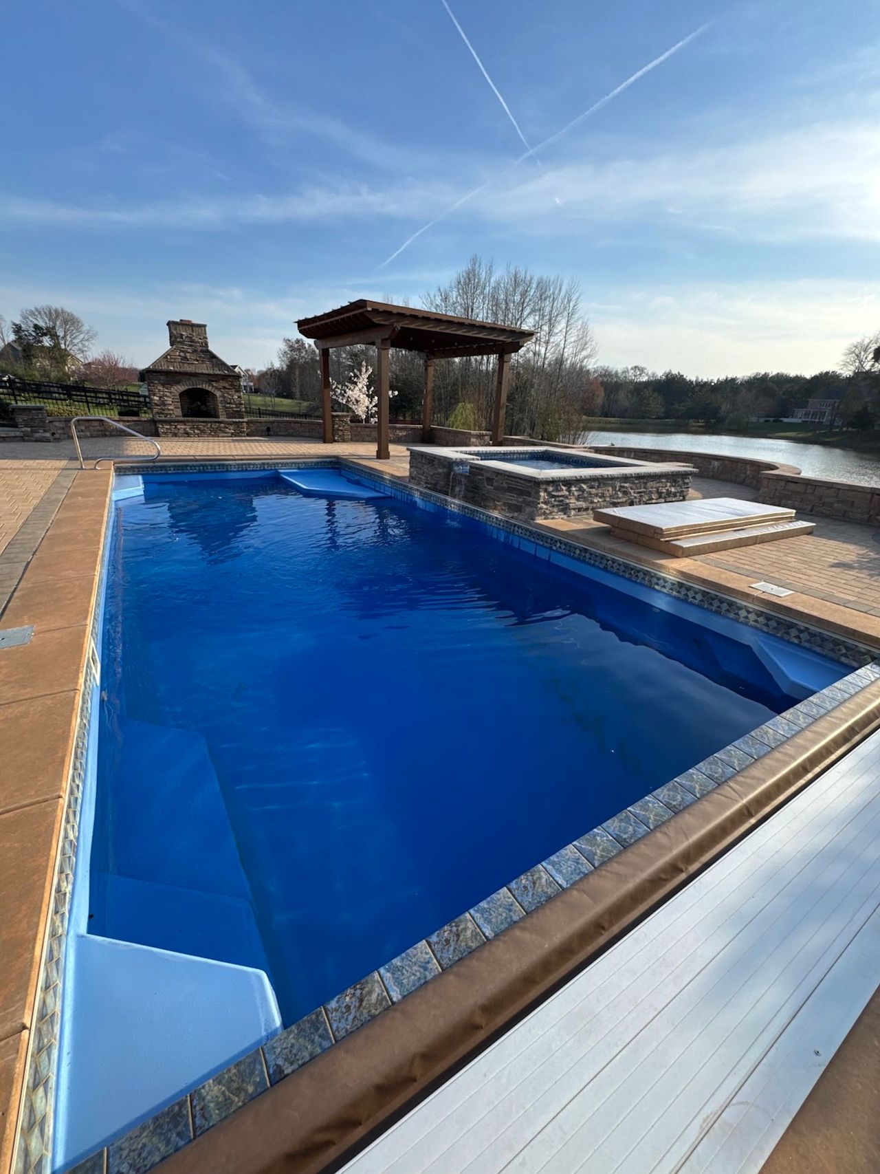 Outdoor infinity pool with bright blue water beside a patio and lakeside view under a clear sky