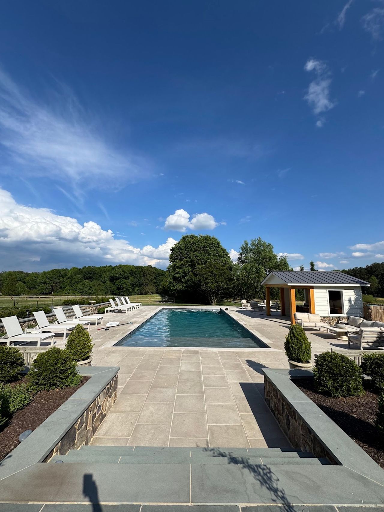 Outdoor pool with stone patio, lounge chairs, and a small pavilion under a blue sky