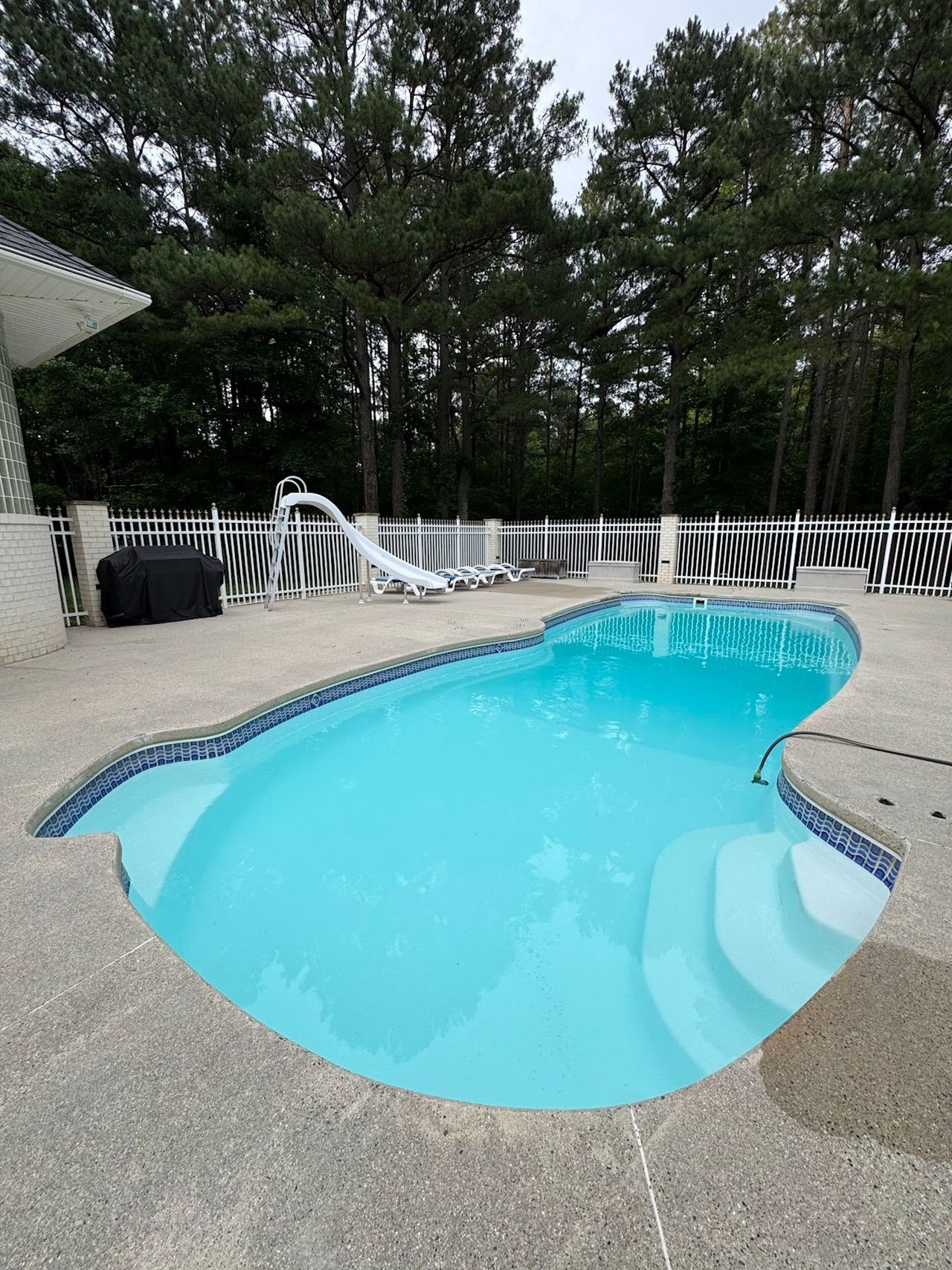 Empty backyard swimming pool with turquoise water and white fence beside trees