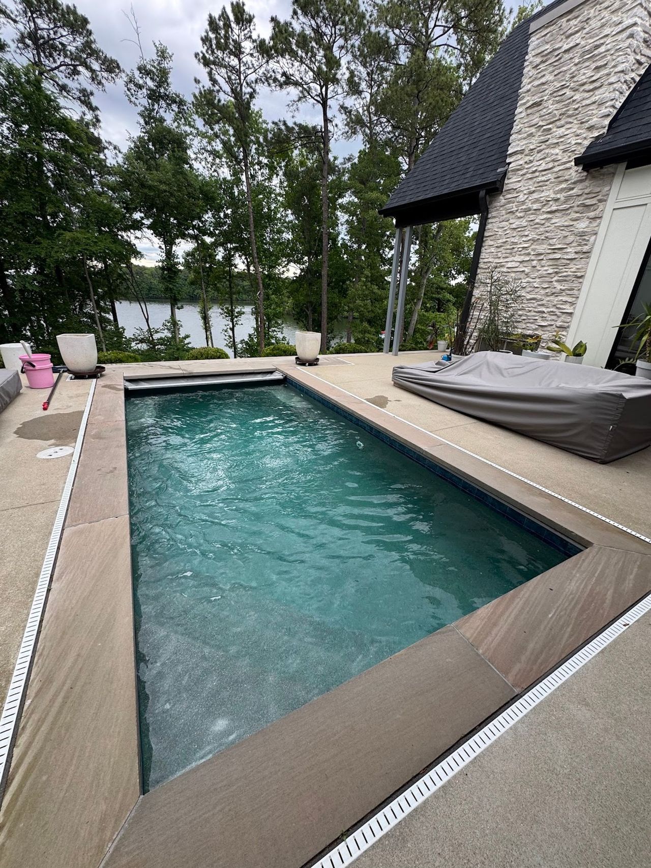 Backyard patio with a rectangular swimming pool beside a stone house and trees.