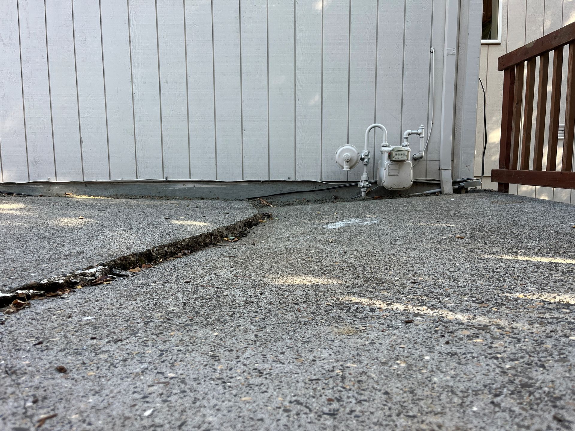 A cracked asphalt patio in front of a white building and gas meter.