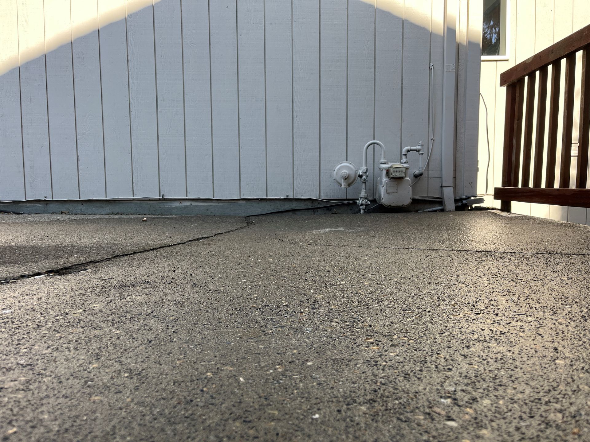 Asphalt patio with crack near white wall, gas meter, and wooden railing.