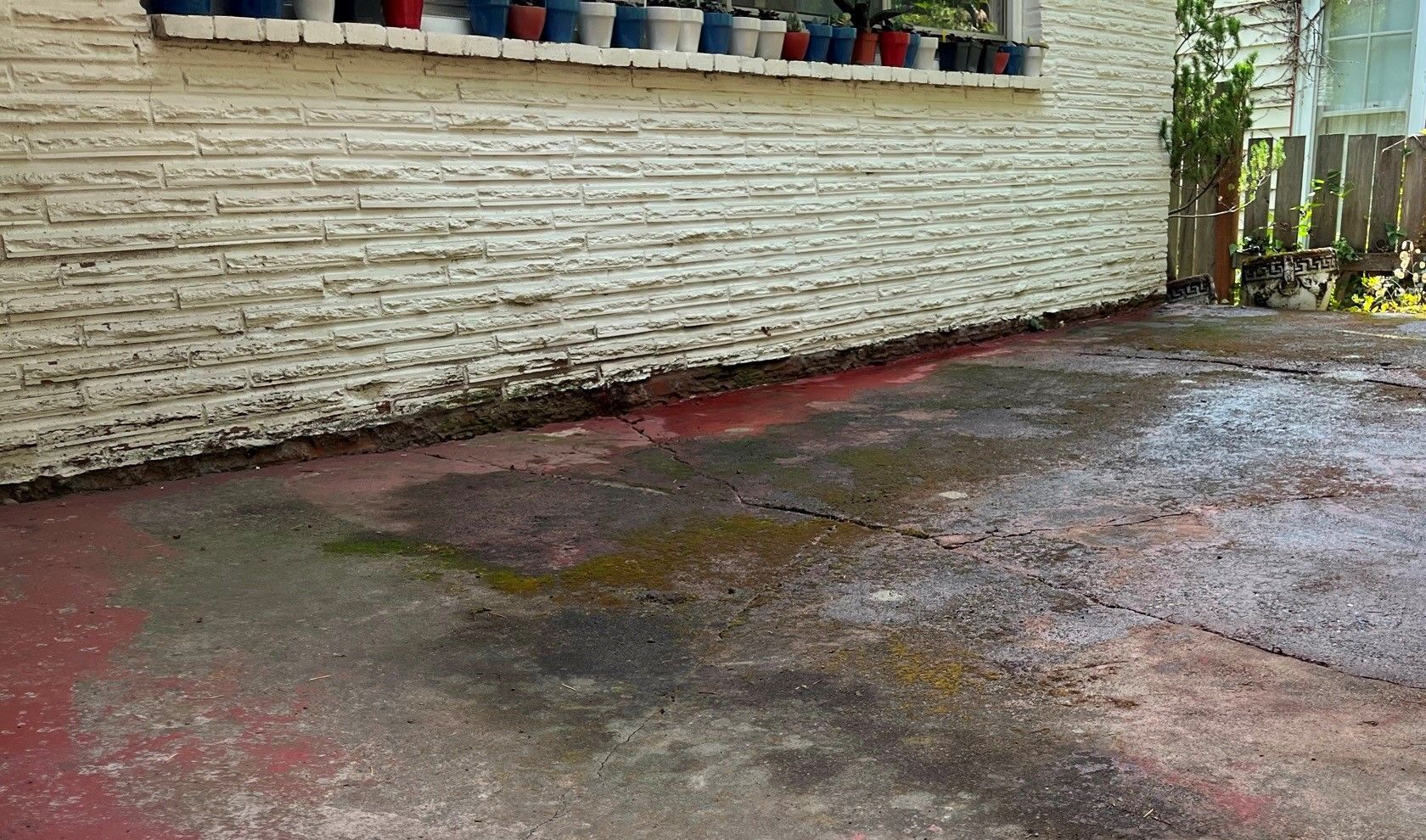 Exterior of a building with a textured white wall, a wet concrete patio with colorful stains, and a window with potted plants.