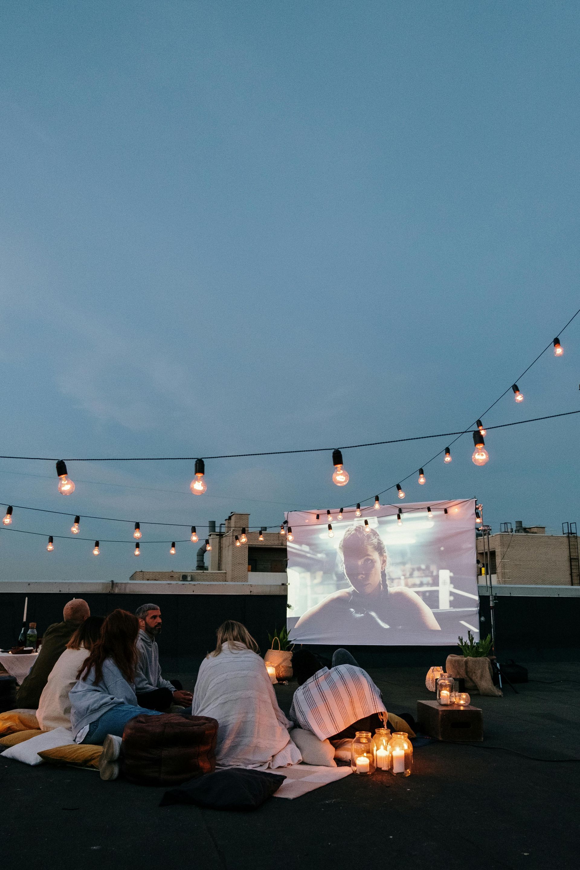 People watch movie on rooftop at night under string lights.