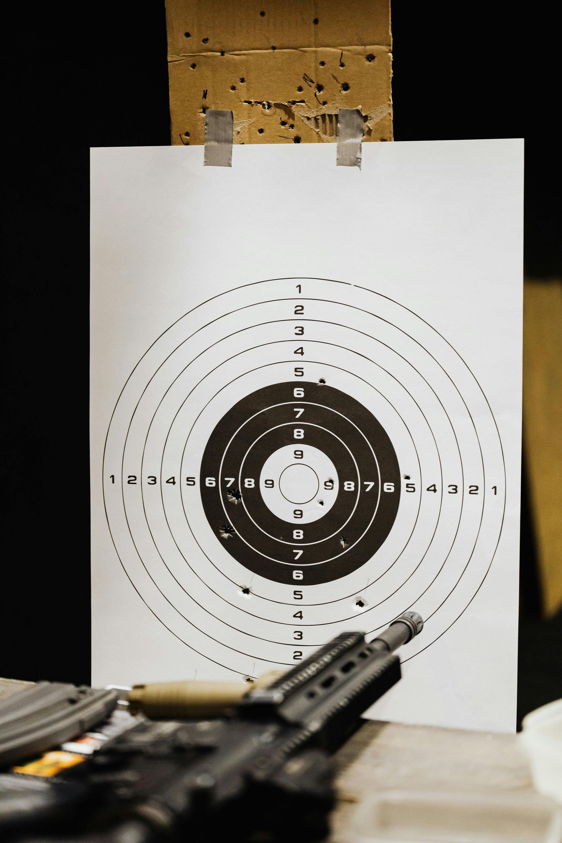 Target paper with bullet holes clipped to a backboard above a rifle on a table.