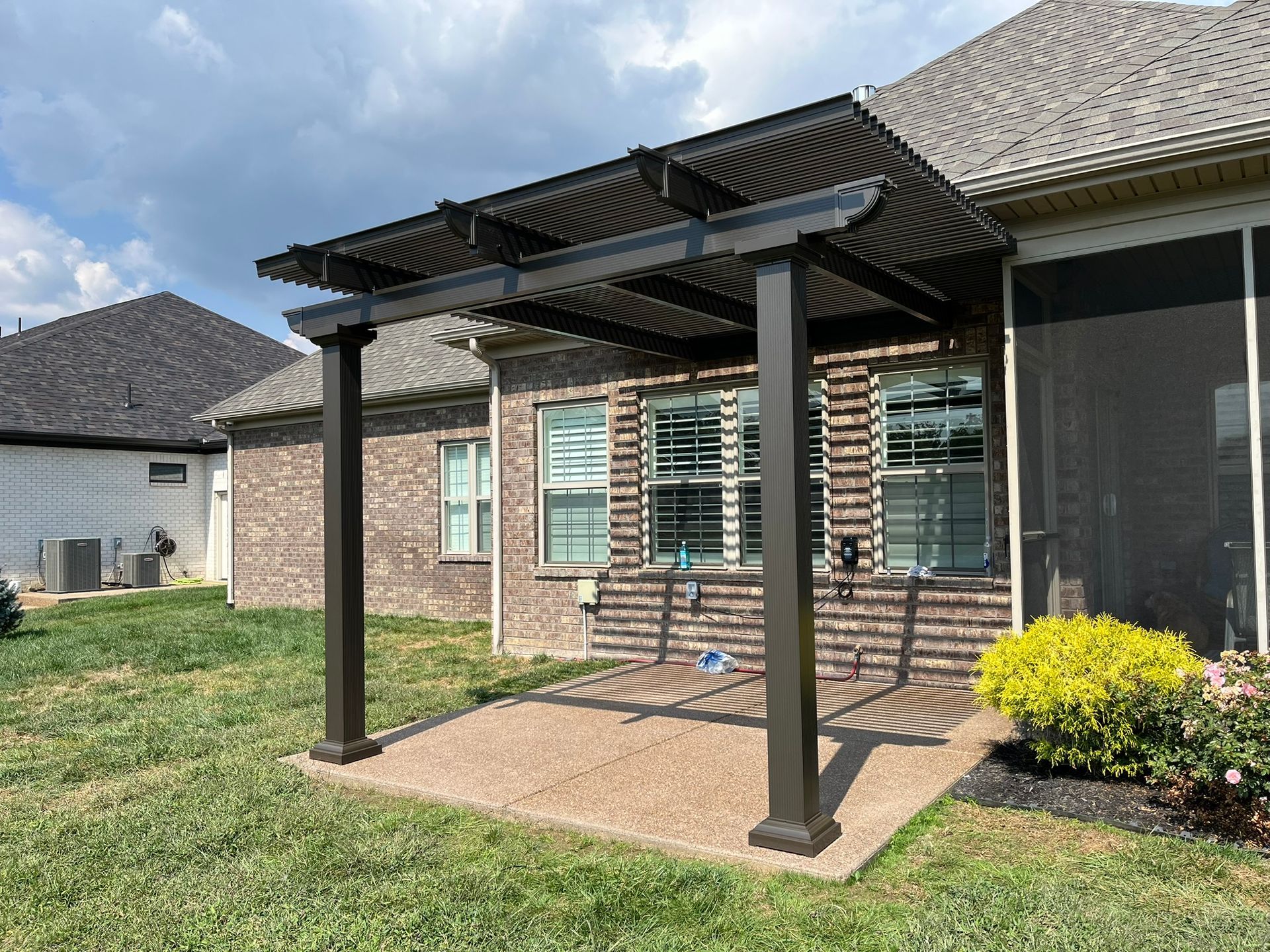 A pergola attached to a brick house, with a concrete patio in a backyard.