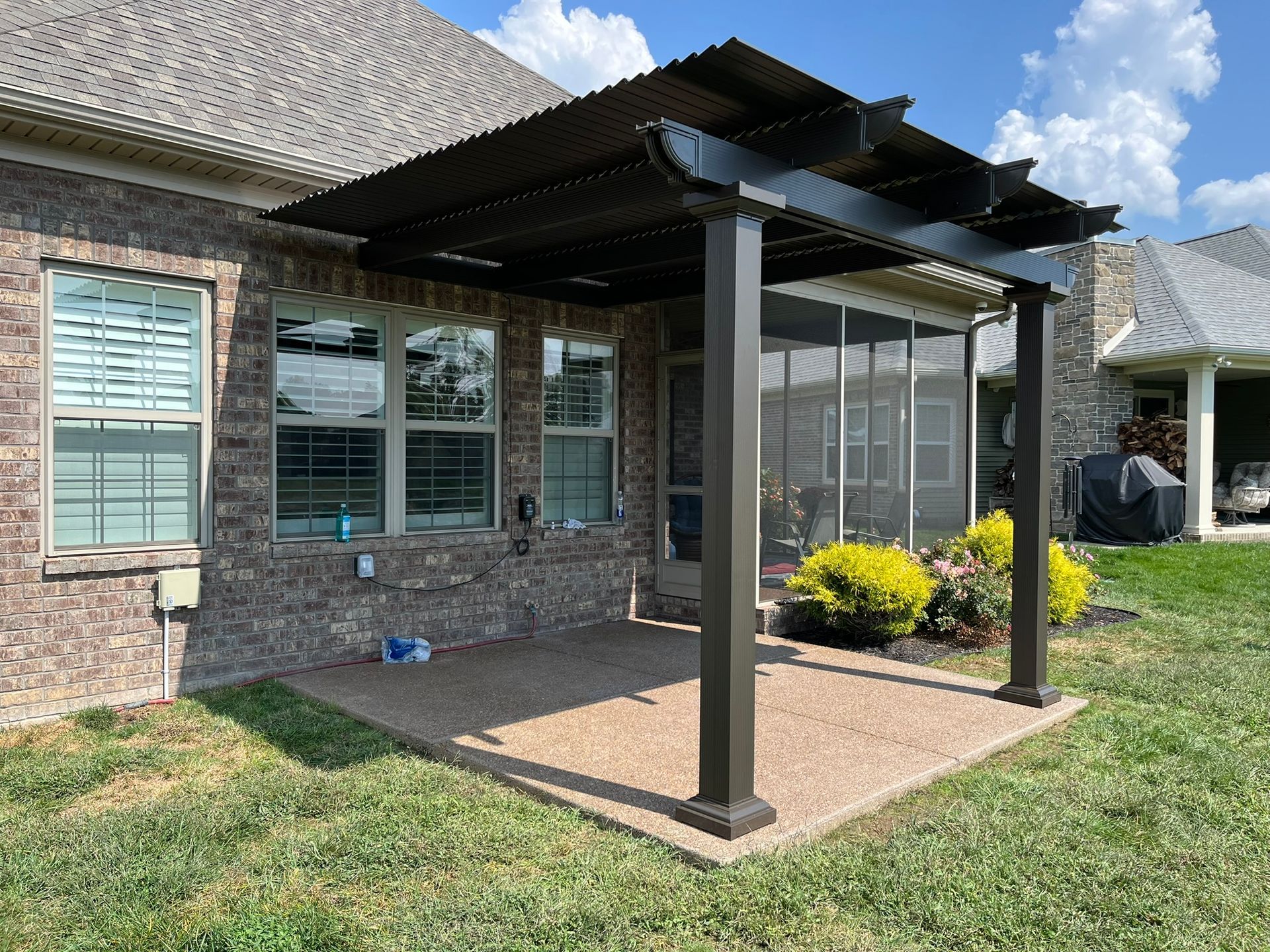 Pergola on a brick patio, with dark brown posts and canopy. Green bushes and lawn surround.