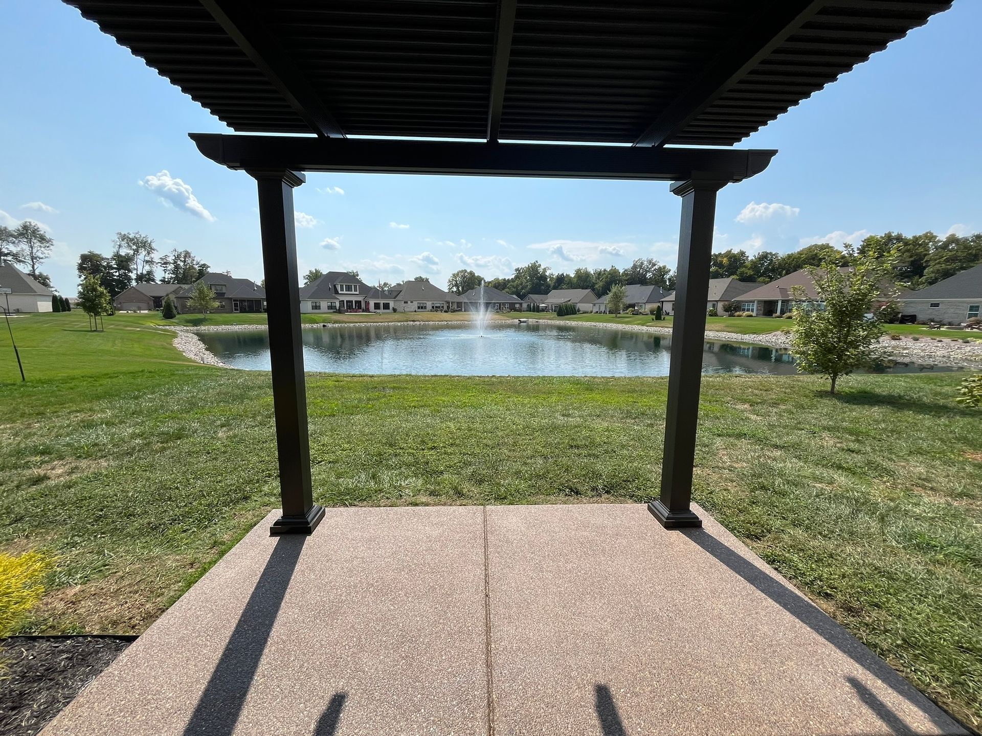A dark pergola frames a pond with a fountain, houses in the distance, and a green lawn