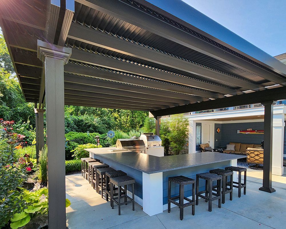 A large outdoor kitchen with a large table and stools under a pergola.