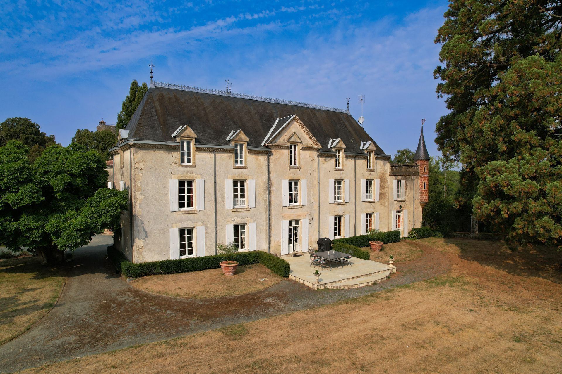 Large stone French chateau with a slate roof, set in a grassy landscape under a blue sky.