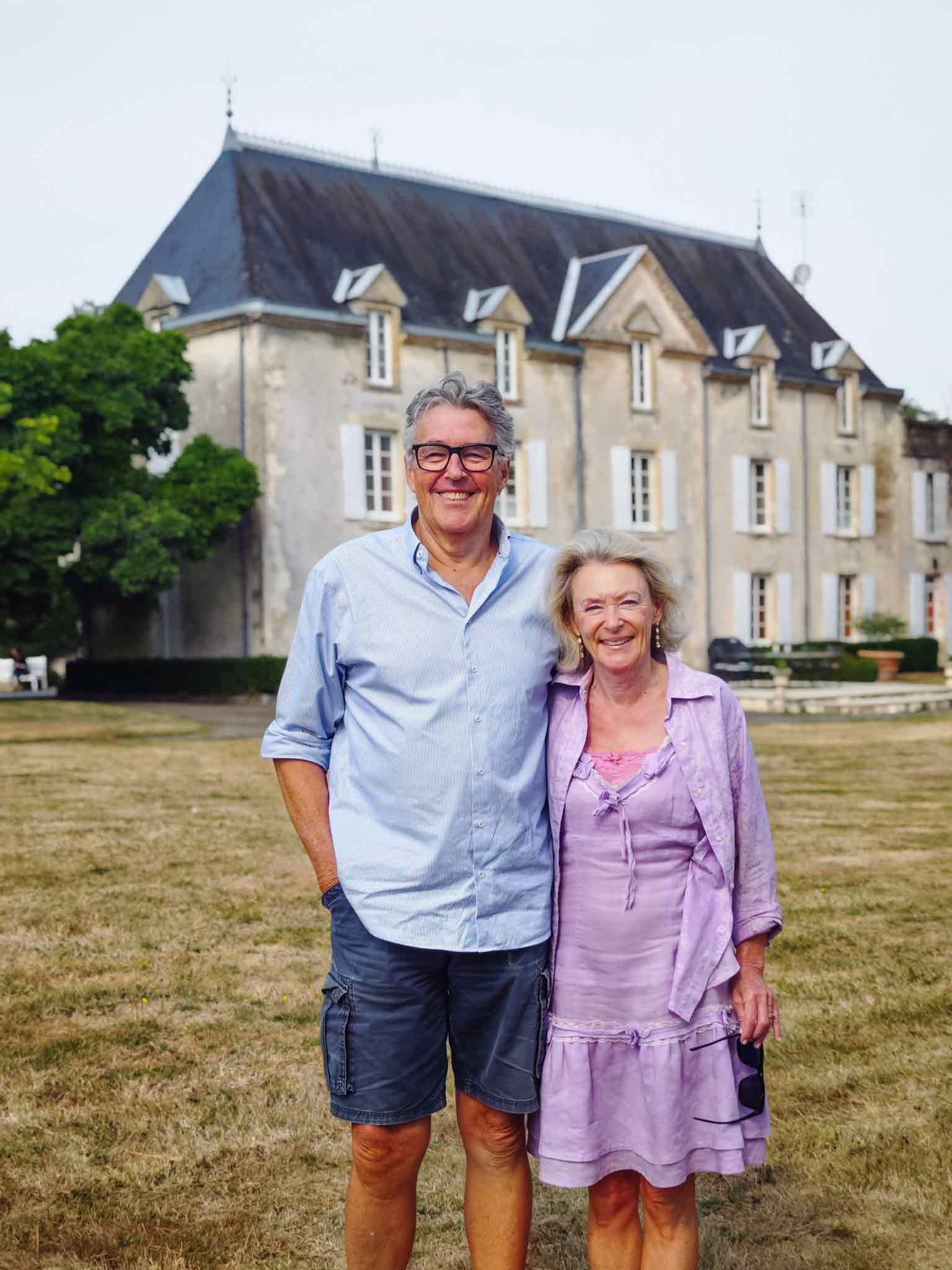 Couple posing in front of a large chateau. The man is wearing a blue shirt and shorts. The woman is wearing a purple dress.