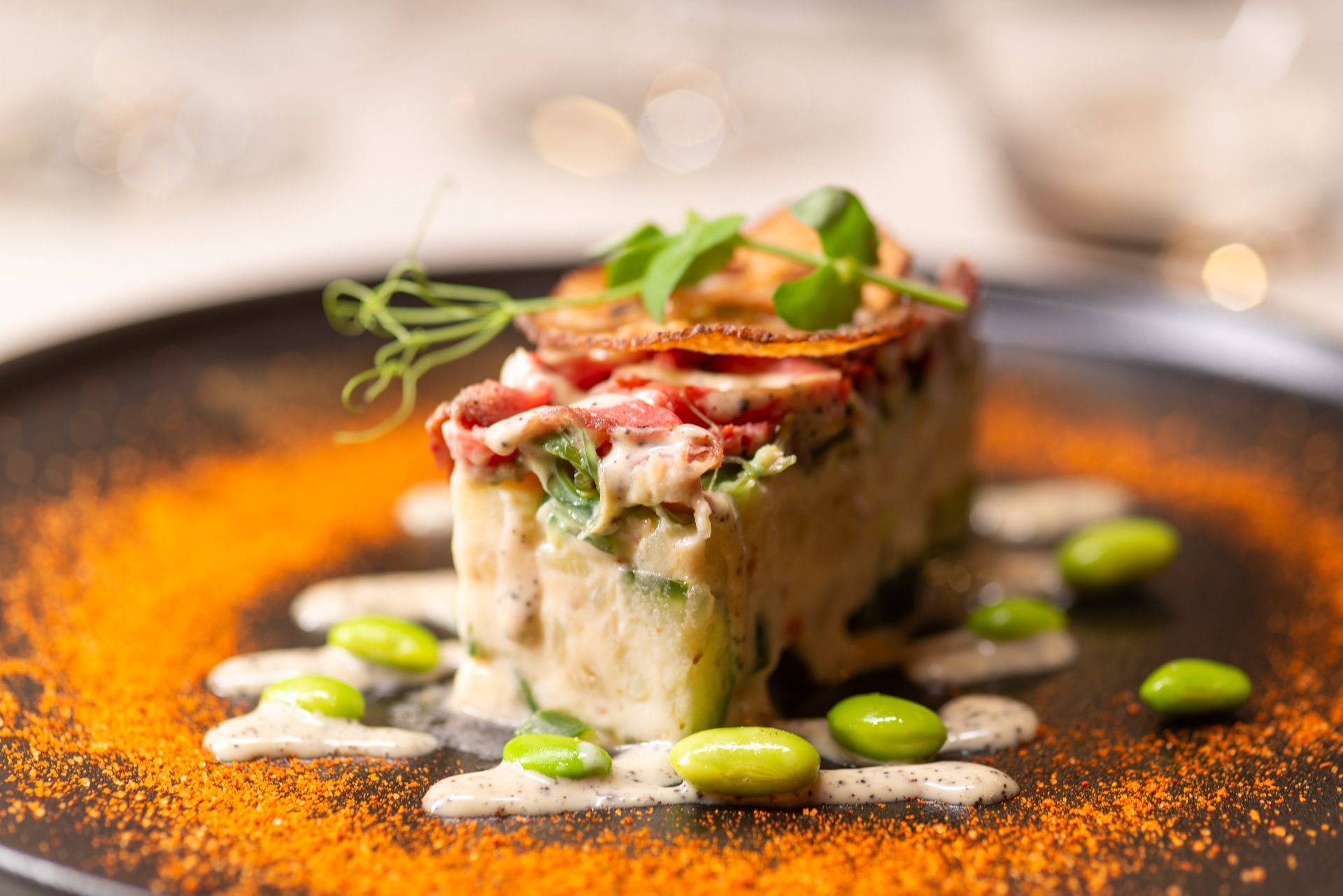 Gourmet-gerecht: kubusvormig gerecht met erwtenspruiten, edamame en sinaasappelpoeder op een zwart bord.