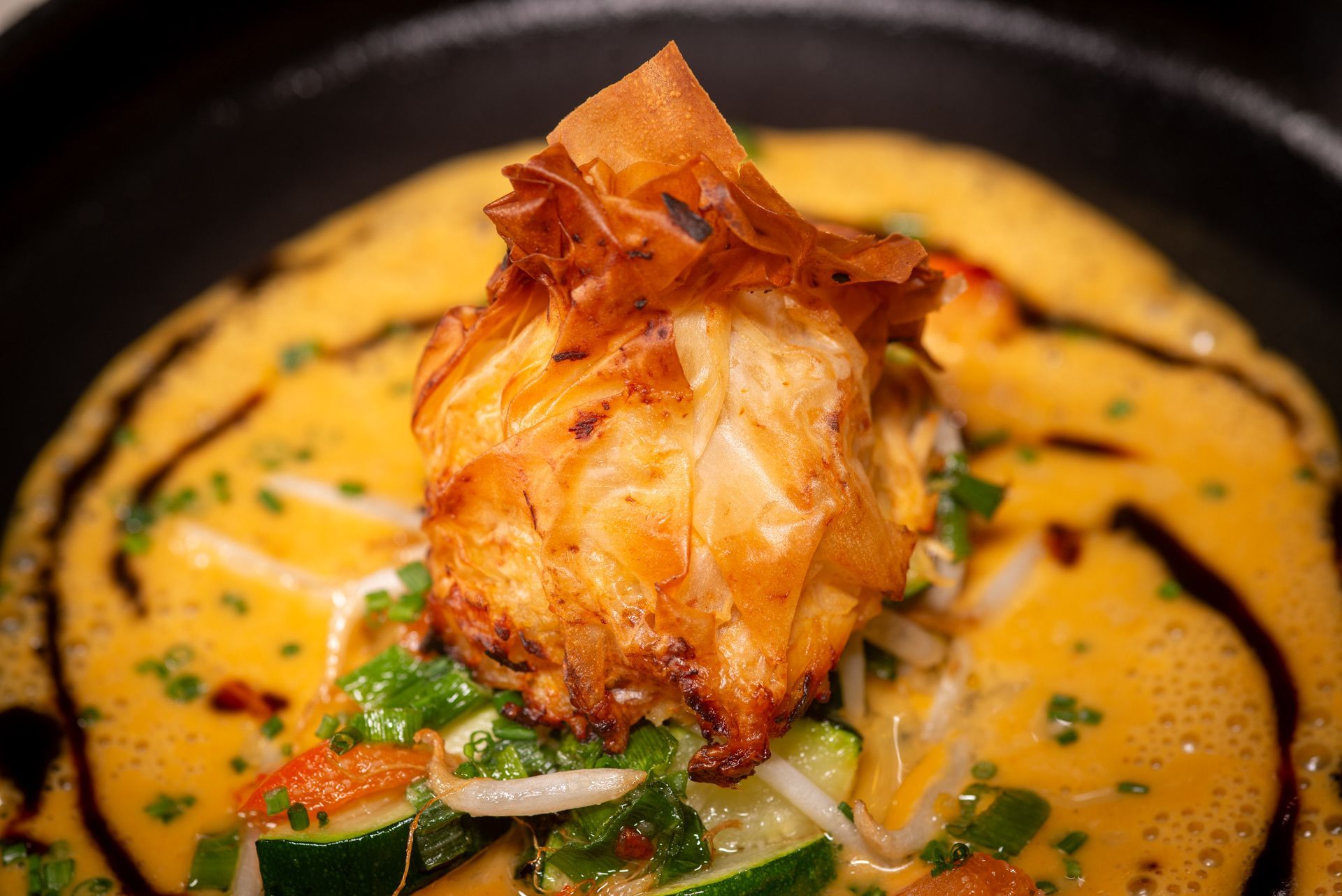 Gefrituurd deeg omwikkeld met gele saus, groene groenten en donkere saus op een zwart bord.