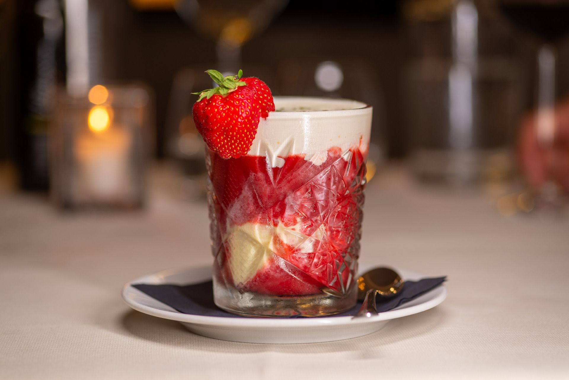 Aardbeiendessert in een glas, met daarop een hele aardbei, geserveerd op een wit bord met een gouden lepel.