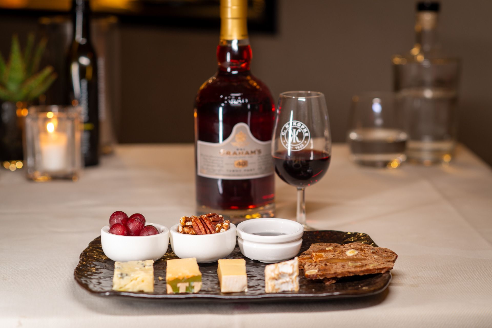 Een fles portwijn met een glas, kaas, druiven, pecannoten en een broodcracker op een dienblad.