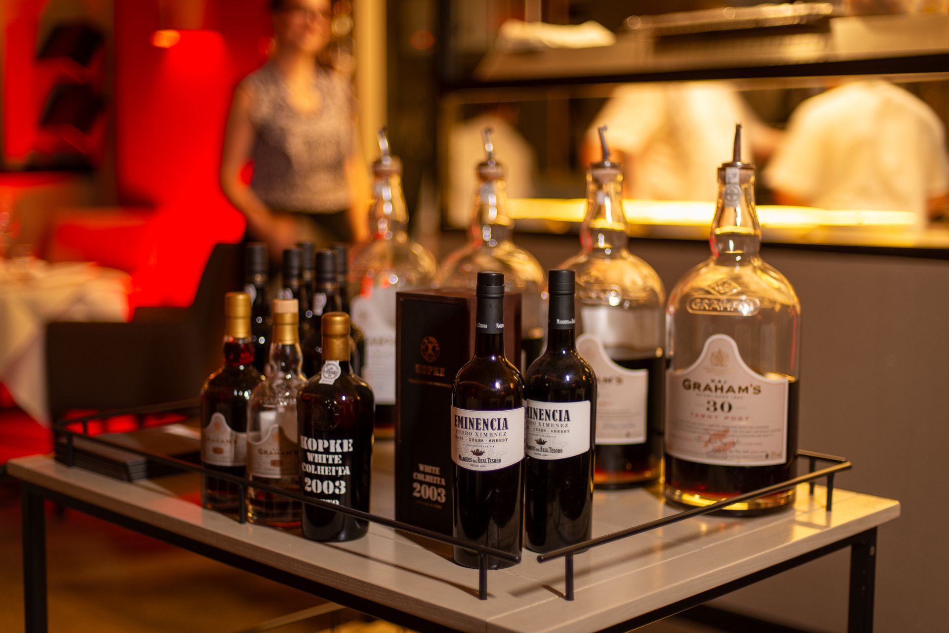 Flessen wijn en sterke drank op een tafel in een restaurant, met mensen in de verte op de achtergrond.