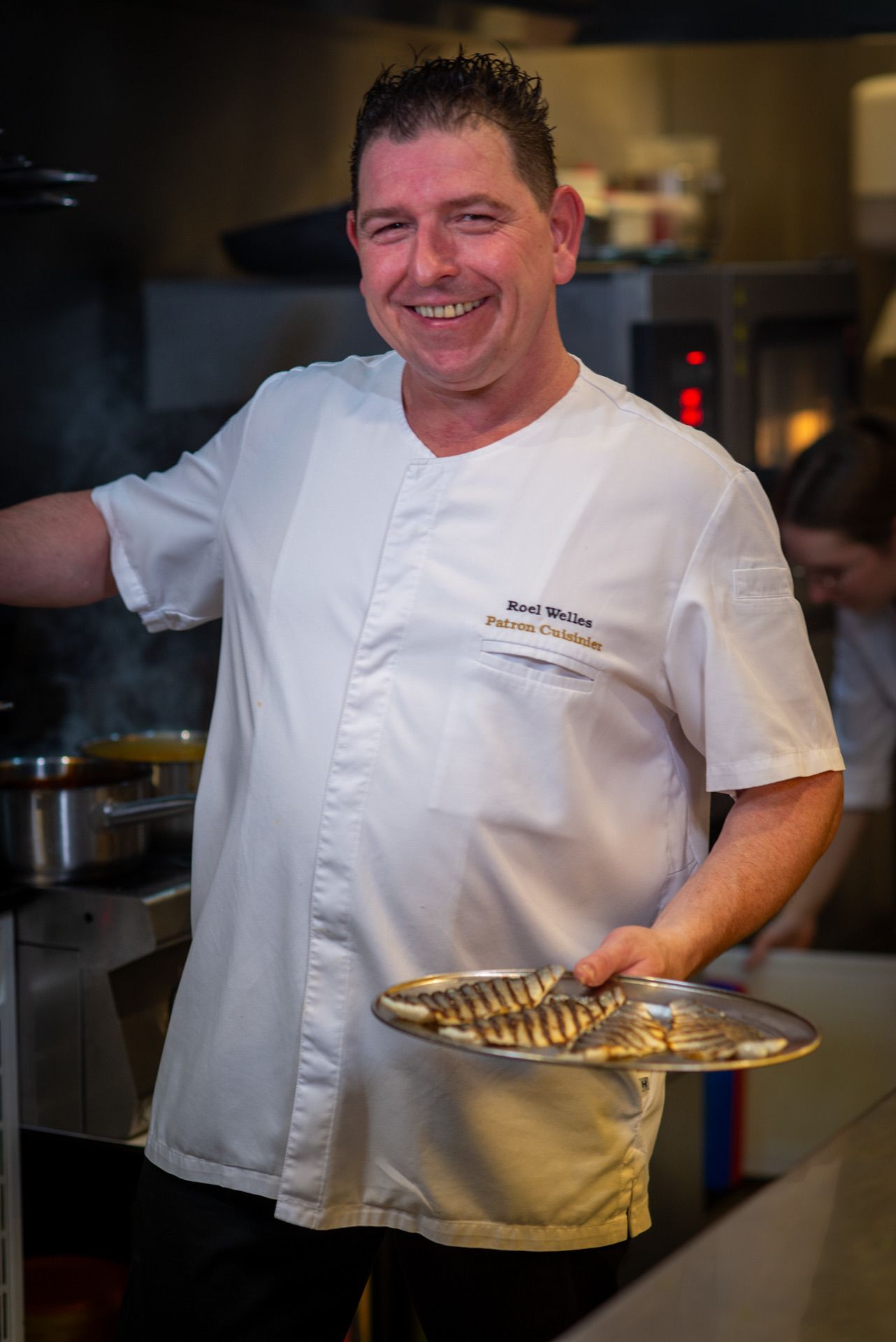Chef in witte jas, met bord in de hand, lachend in de keuken.