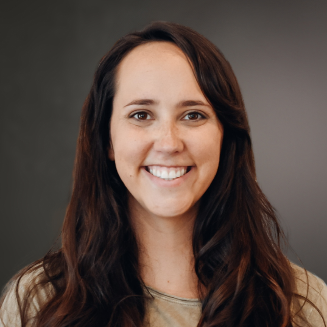 Woman with long brown hair smiling at the camera, wearing a light-colored top, against a gray background.