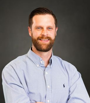 A man with a beard is wearing a blue shirt and smiling with his arms crossed.