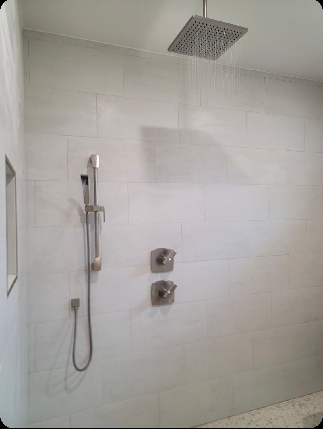 Shower with square rain showerhead, handheld shower, and two silver faucet handles on light gray tiled walls.