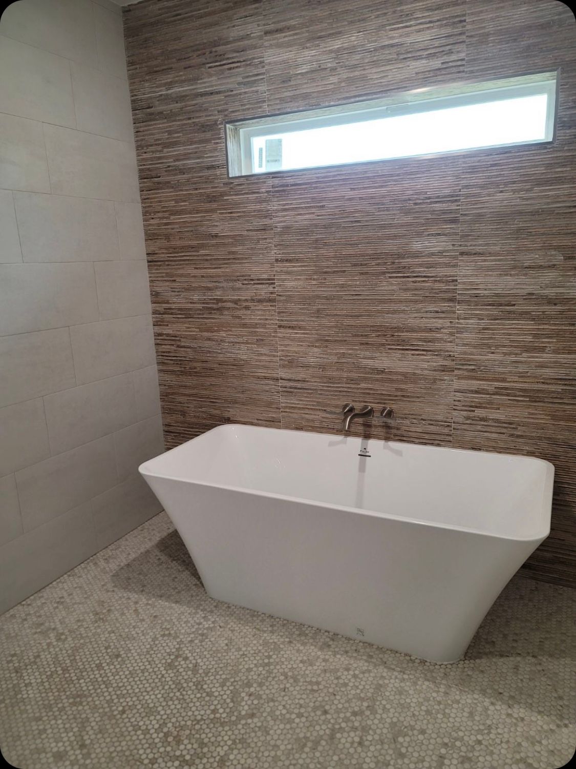 Freestanding white bathtub in a bathroom with mosaic floor and brown textured accent wall.