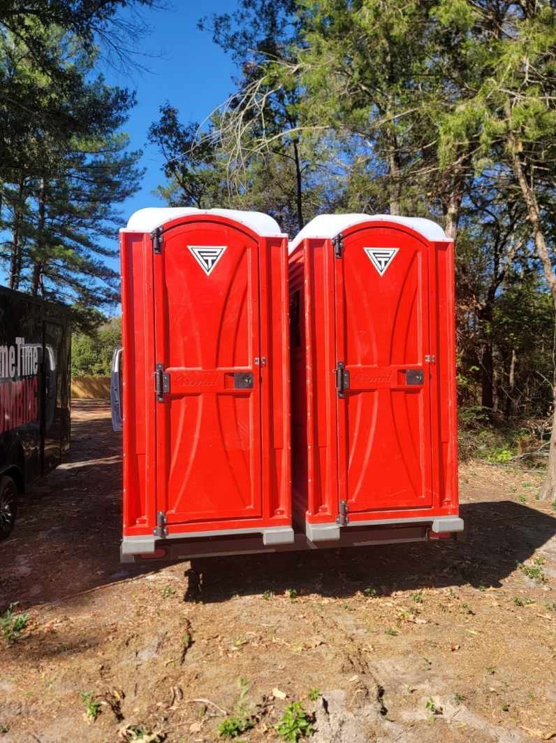 Two bright red portable toilets in a wooded area, with white tops and logos.