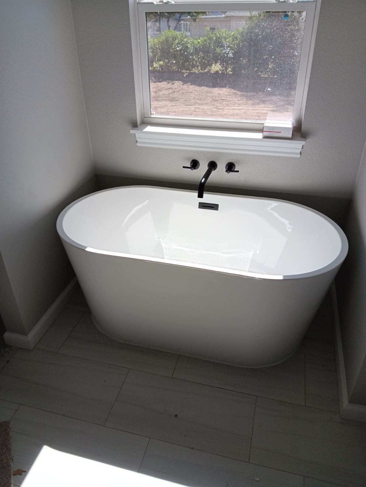 Oval white bathtub in a bathroom, beneath a window. Black faucet on the wall. Light-colored tile floor.