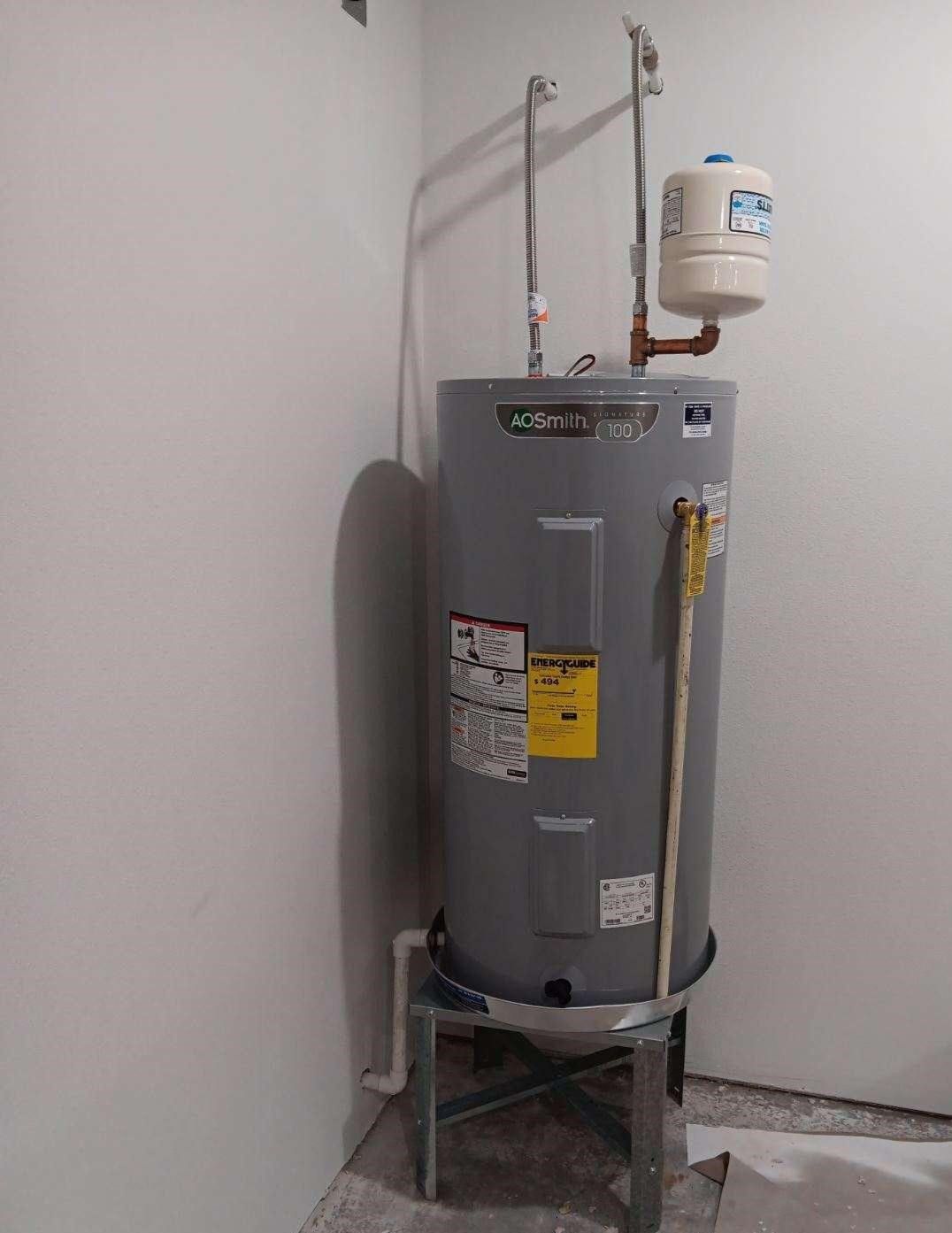 Gray water heater in a corner, with pipes, a white expansion tank, and a metal stand.