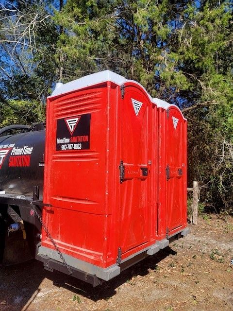 Two red portable toilets on a trailer, parked outdoors near trees.