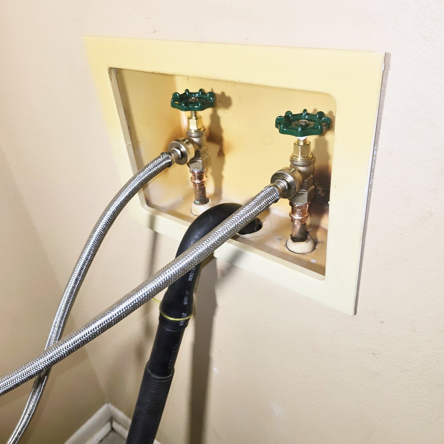 White wall with utility box containing two faucets and drainpipe, connected by silver hoses.