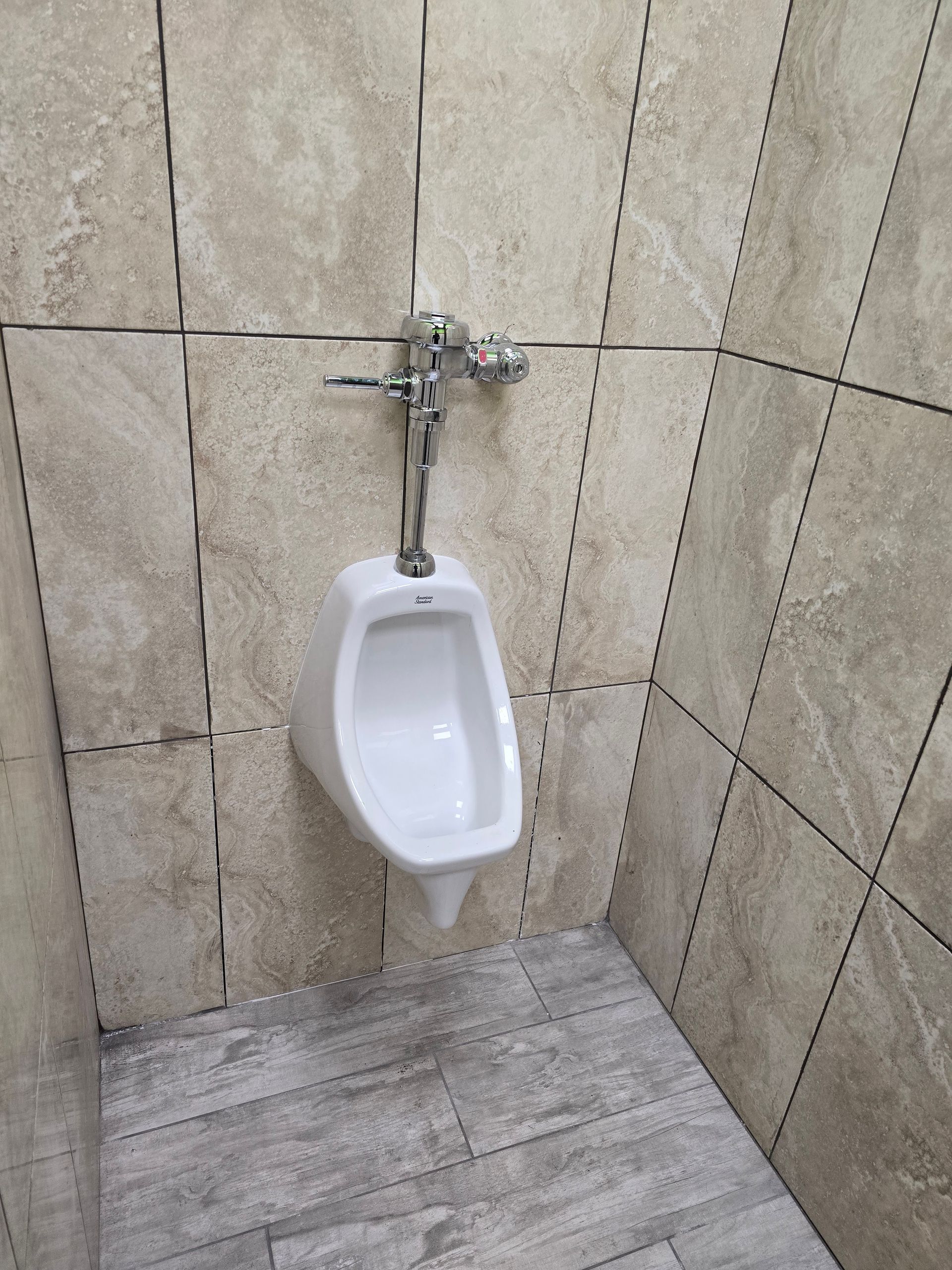 Urinal mounted on tan tiled wall in a restroom with gray tile flooring.