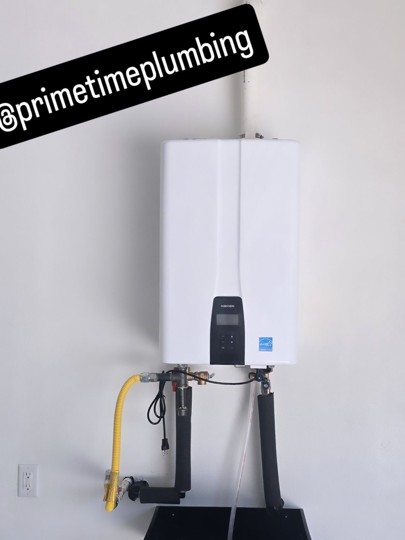 White tankless water heater on white wall with black pipes and gas line.