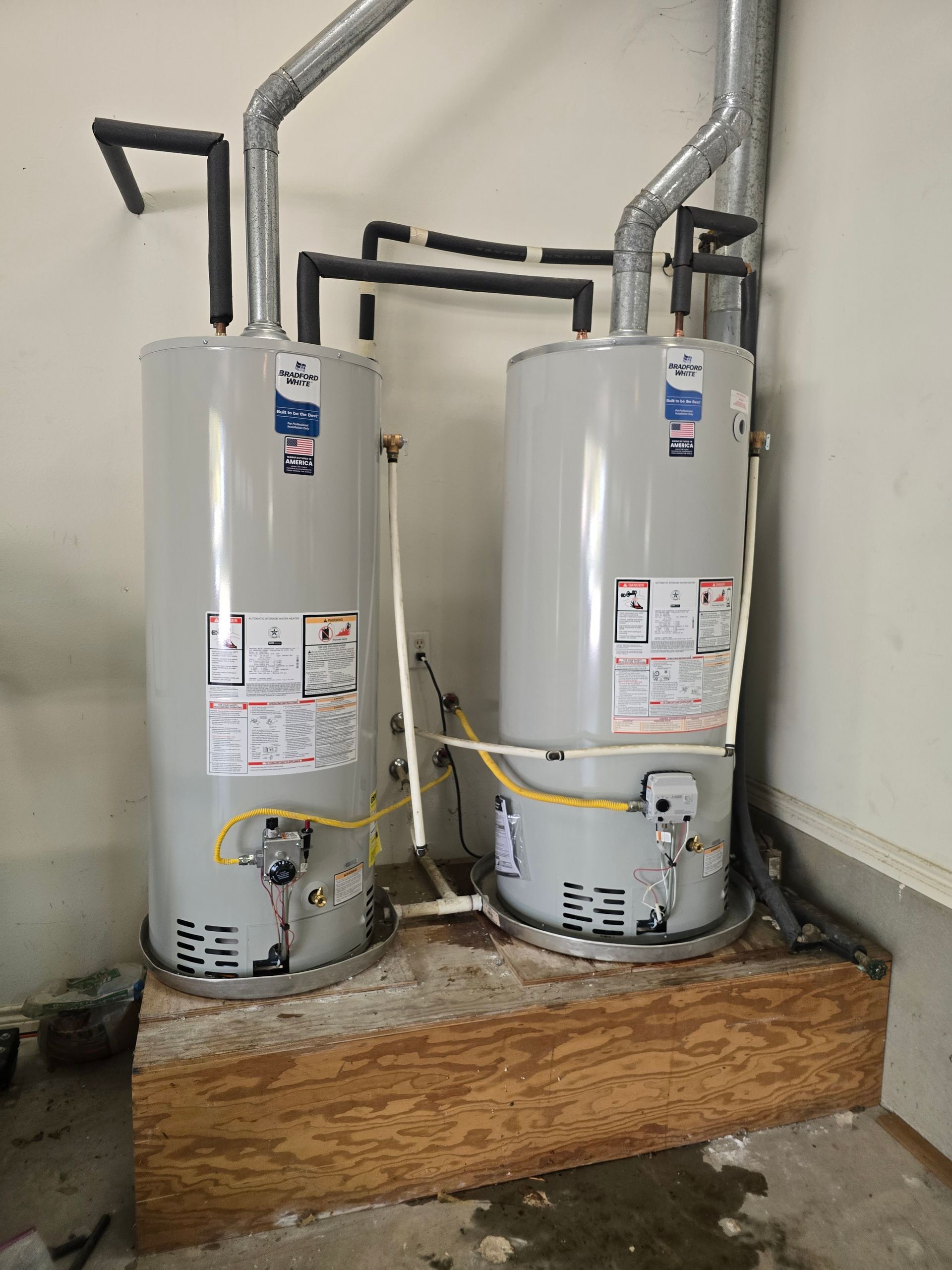 Two tall gray water heaters side-by-side with connected pipes and vents, mounted on a wooden platform against a wall.