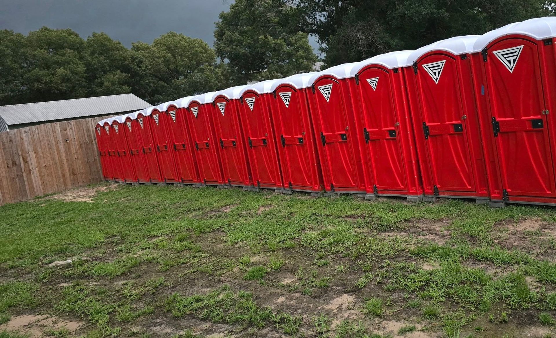 Red portable toilets lined up on grass next to a wooden fence and trees.