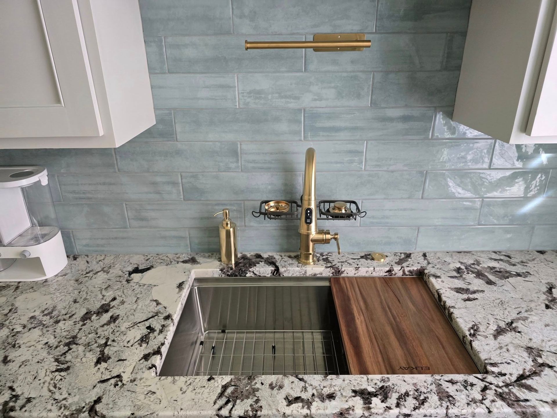 Kitchen sink with gold faucet, cutting board, and blue tile backsplash. White cabinets and granite countertop.