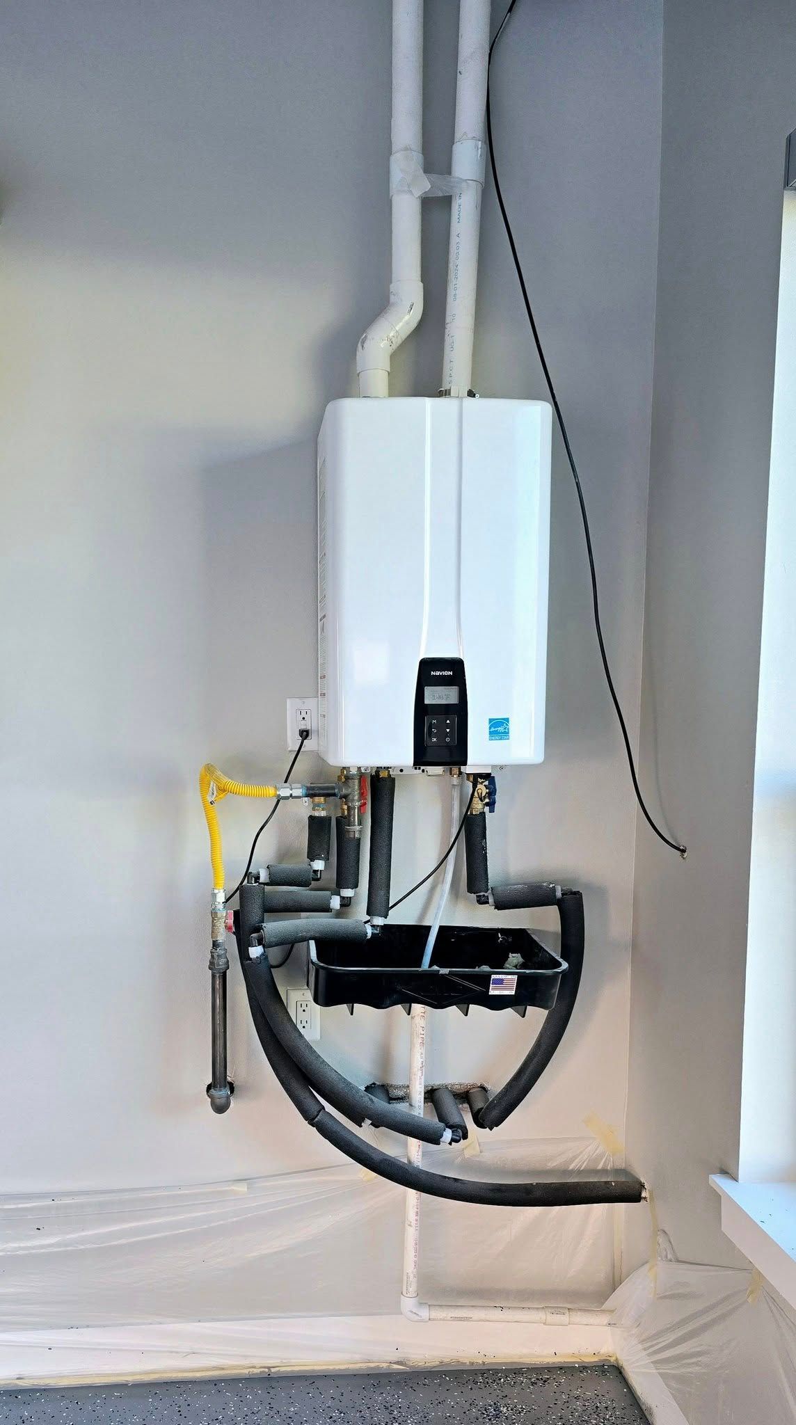 White tankless water heater mounted on a light gray wall. Pipes and wiring visible.