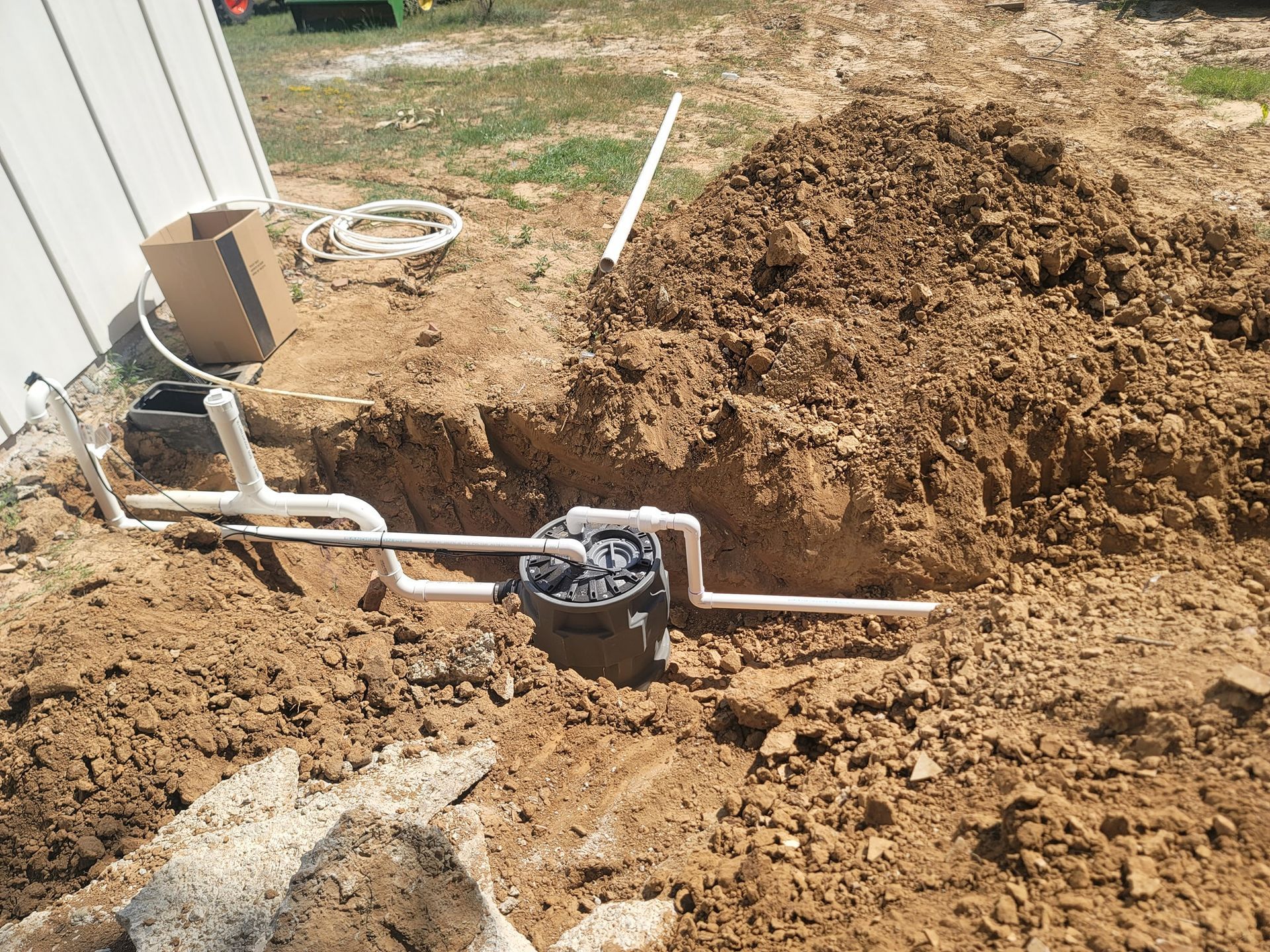 Pipes connected to a black box in a dirt trench, near a white wall and a pile of dirt.