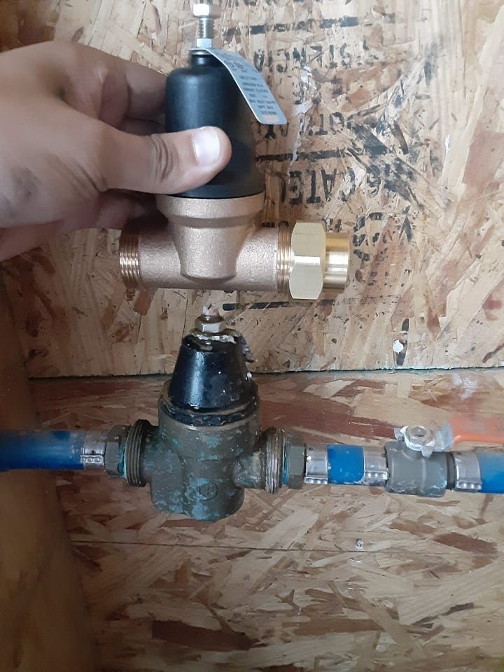 Hand holding a black and gold pressure regulator above an older brass one, attached to blue and metal pipes, against wood.